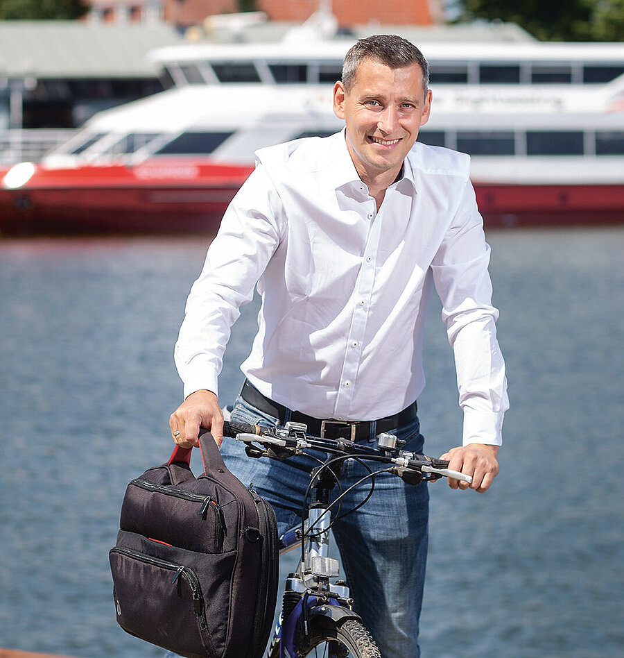 Christian Stolt, Pflegegutachter beim Medizinischen Dienst Mecklenburg- Vorpommern (auf dem Fahrrad, im Hintergrund ein Schiff auf dem Wasser)